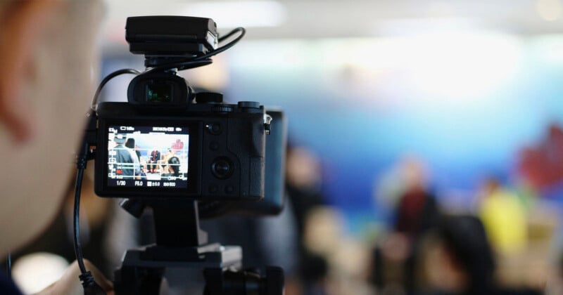 A close-up of a digital camera on a tripod filming a group of people in a brightly lit, blurred background setting, possibly during an interview or discussion.