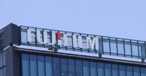 The Fujifilm logo in white lettering with a red dot above the "i" is mounted on the roof of a modern glass-paneled building against a clear blue sky.