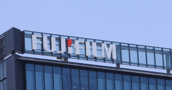 The Fujifilm logo in white lettering with a red dot above the "i" is mounted on the roof of a modern glass-paneled building against a clear blue sky.