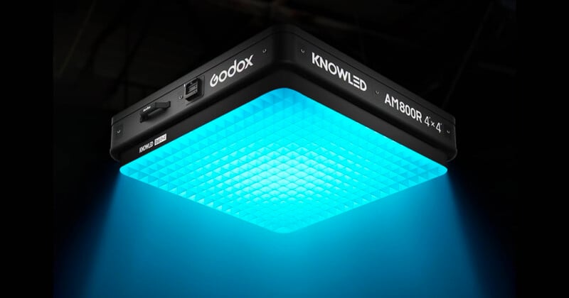 A square Godox KNOWLED AM1800R LED light panel emits a bright blue glow downward, illuminating the area below in a dark environment.