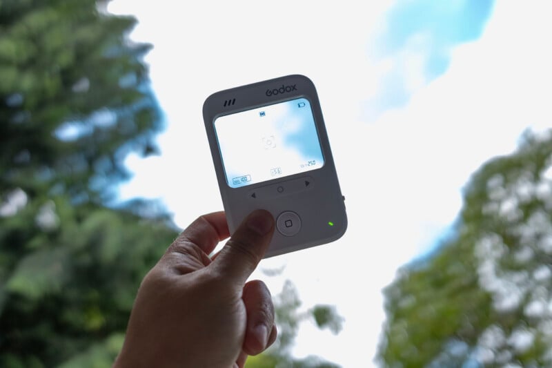 A hand holds up a Godox device with a digital screen outdoors. Trees and blue sky with clouds are visible in the background.