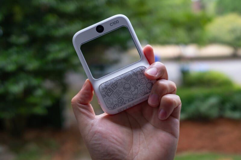A hand holds a small white device labeled "C100" with a clear rectangular screen and a patterned lower section, against a blurred outdoor background with greenery.