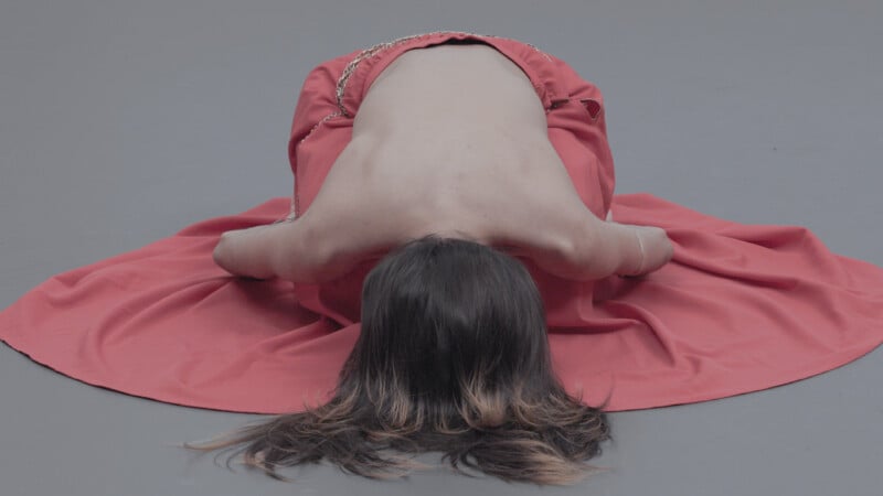 A person with long hair, bare back, and covered in a red cloth, kneels and bows forward on a gray surface, arms bent and head down.