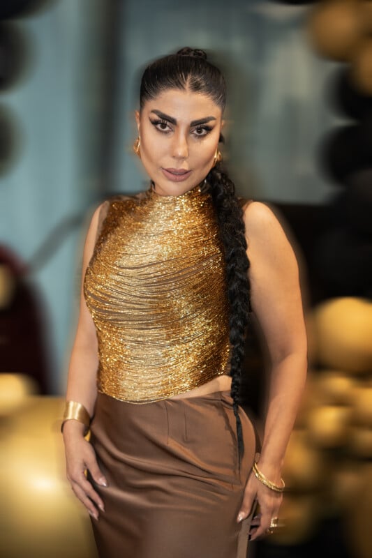 Woman wearing a gold, sleeveless top and brown skirt, with a long braided ponytail, poses confidently in front of a blurred background with black and gold balloons.