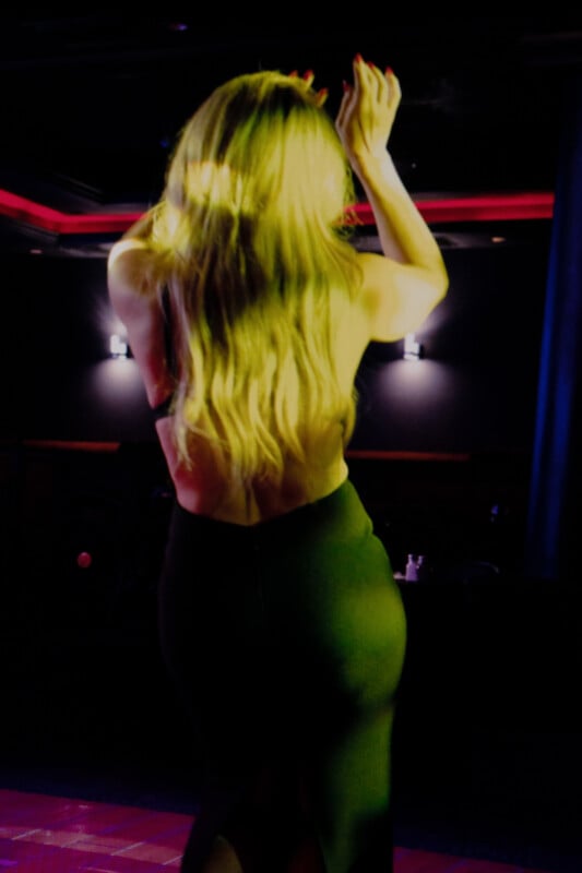 A woman with long, wavy blonde hair stands with her back to the camera in a dimly lit room, wearing a black backless dress and raising her hands above her head.