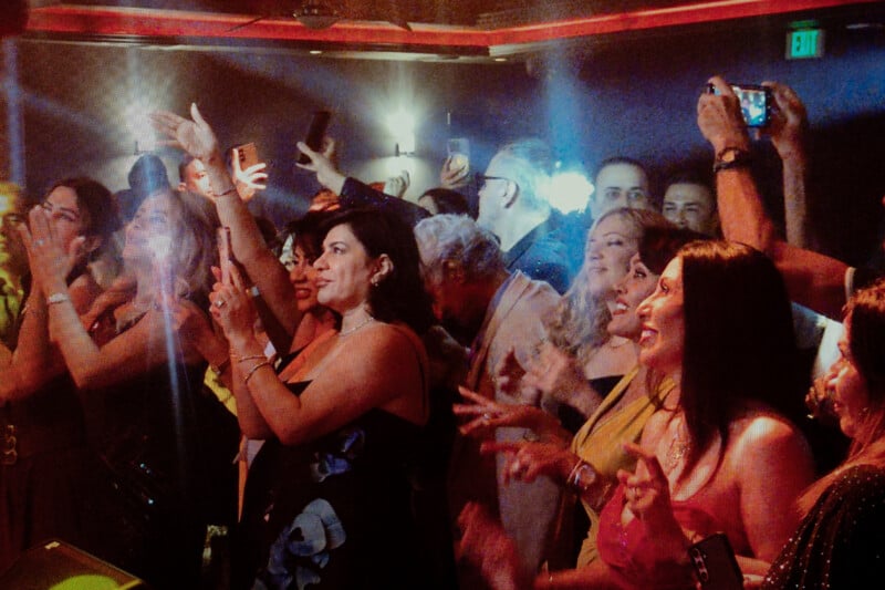 A crowd of people in evening attire cheer, laugh, and take photos with their phones at a lively indoor event, surrounded by warm lighting and excitement.