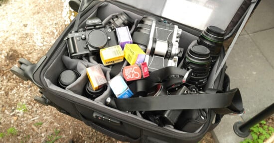 A camera bag filled with DSLR cameras, lenses, and several boxes of film in various colors, resting outdoors on the ground next to grass and pavement.
