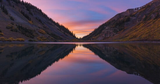 A serene mountain lake reflects vibrant pink, purple, and orange hues from a dramatic sunset sky, nestled between two forested mountain slopes.