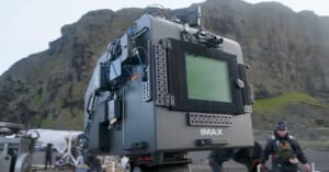 A large IMAX camera is set up outdoors with rocky hills in the background. Film crew members and equipment are visible around the scene.