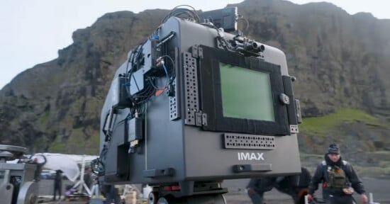A large IMAX camera is set up outdoors with rocky hills in the background. Film crew members and equipment are visible around the scene.