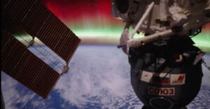 A view from the International Space Station shows a Russian Soyuz spacecraft and solar panels above Earth, with a glowing green and red aurora visible over the planet’s horizon.