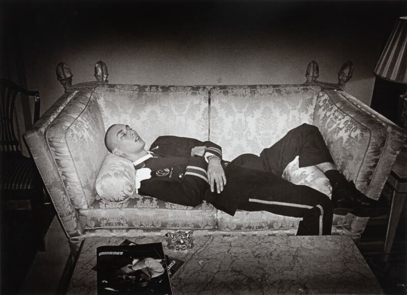 A person in a uniform lies asleep on a patterned sofa, hands crossed over the chest. A magazine and ashtray sit on a marble coffee table in the foreground. The scene is dimly lit.