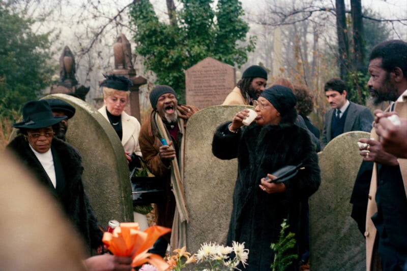 People gathered at a cemetery, some in black coats and hats, some appearing emotional, standing near gravestones with flowers in the foreground.