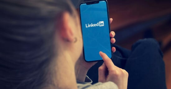 A person holds a smartphone displaying the LinkedIn logo on its screen while touching the display with their index finger.