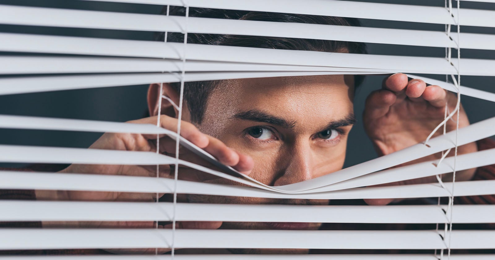 A person peers through open blinds with both hands, looking intently outside.
