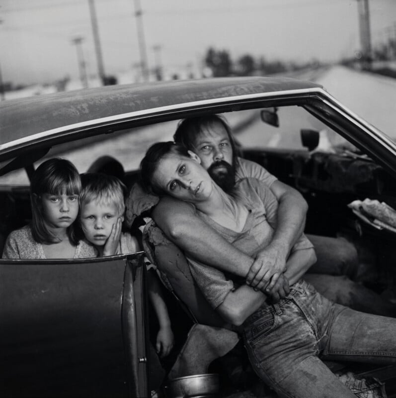 A black-and-white portrait of a family of four sitting inside a car, with a man embracing a woman while two young children look out from the back seat.