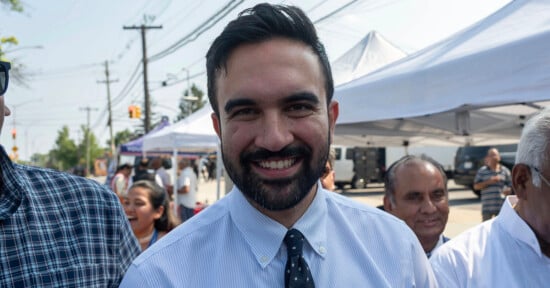 A man with dark hair and a beard, smiling, stands outdoors at an event with people and tents in the background.