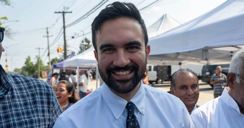 A man with dark hair and a beard, smiling, stands outdoors at an event with people and tents in the background.