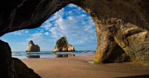 A view from inside a dark rock cave looking out onto a sandy beach with two large rock formations rising from the ocean.