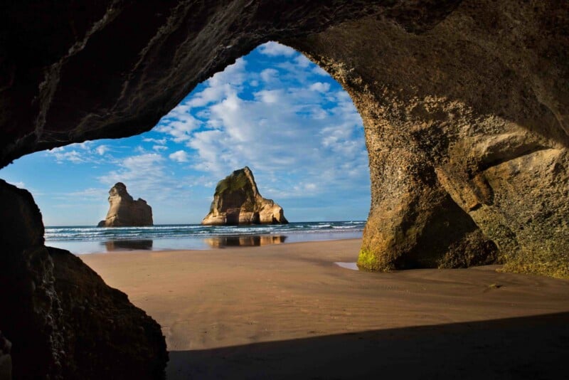 A view from inside a dark rock cave looking out onto a sandy beach with two large rock formations rising from the ocean.