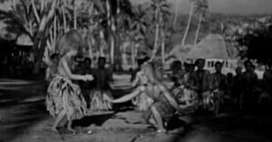 Two people in traditional attire dance outdoors on a sandy area, surrounded by a group of onlookers with palm trees and huts in the background. The image is in black and white.