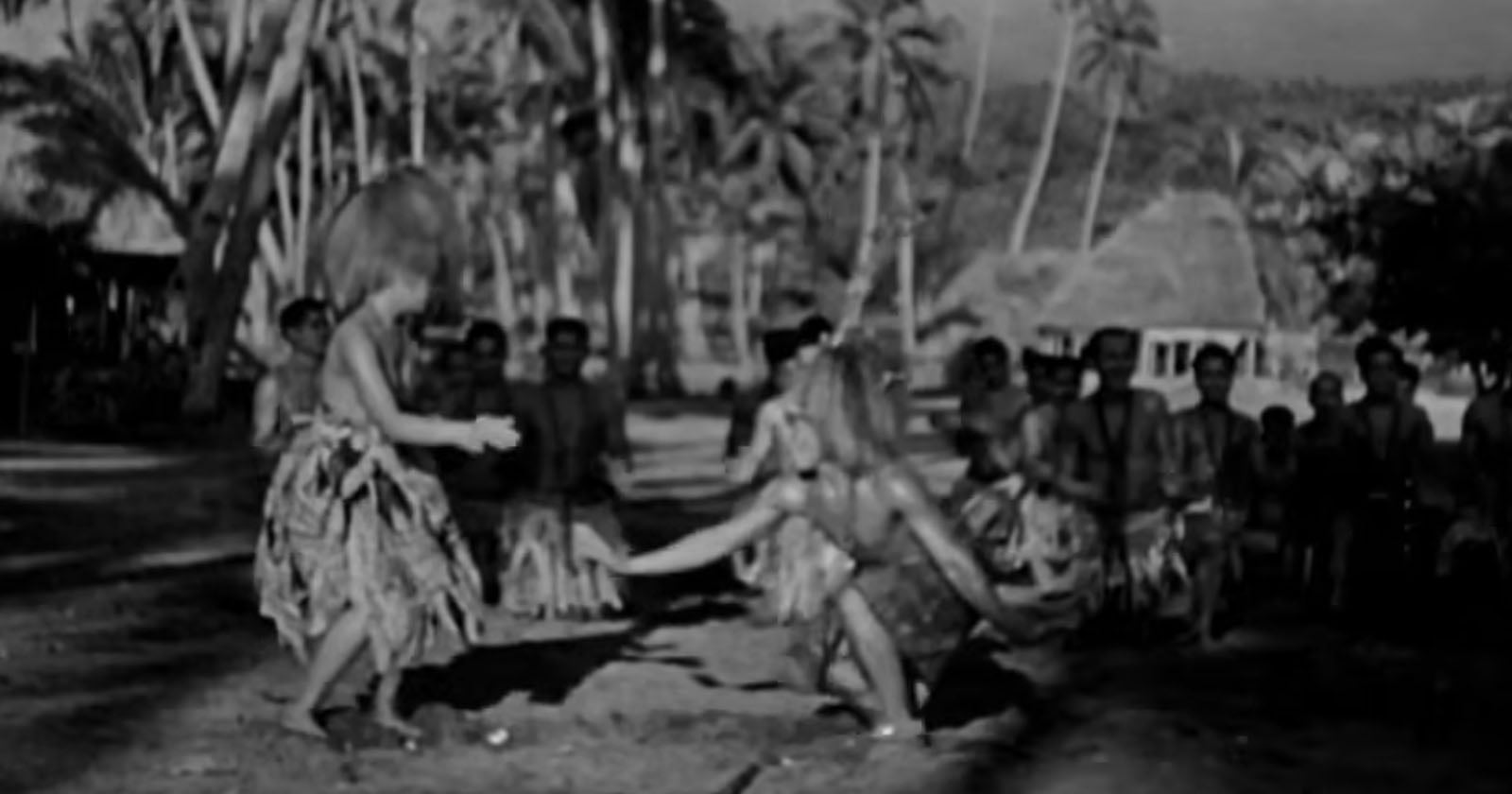 Two people in traditional attire dance outdoors on a sandy area, surrounded by a group of onlookers with palm trees and huts in the background. The image is in black and white.
