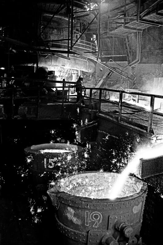 Molten metal pours into a large industrial vat, creating sparks. A worker stands in the background near railings in a dark, gritty factory setting filled with heavy machinery and pipes.