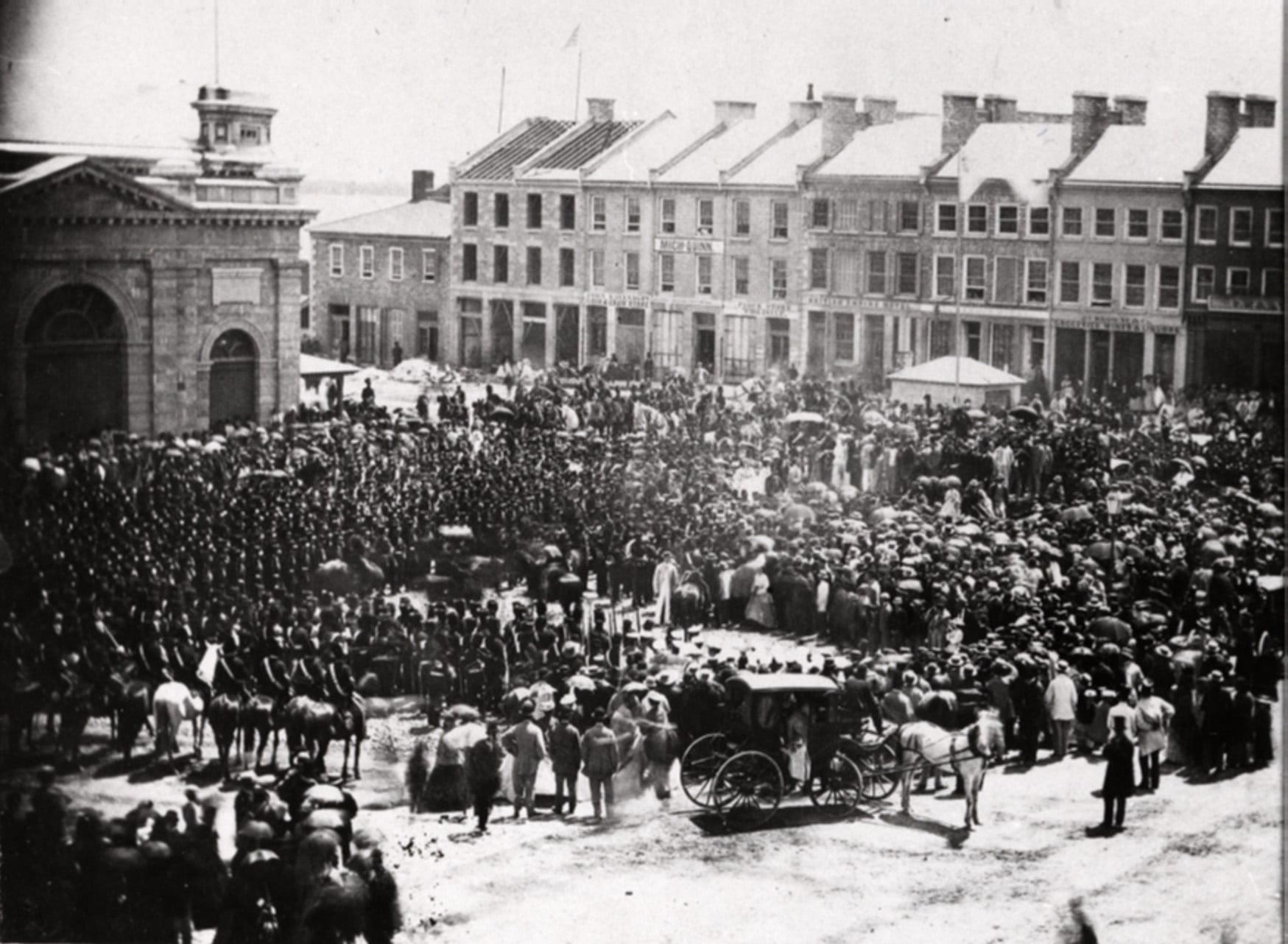 A historic black-and-white photo of a large crowd gathered in a town square, with rows of soldiers, spectators, horse-drawn carriages, and 19th-century buildings in the background.