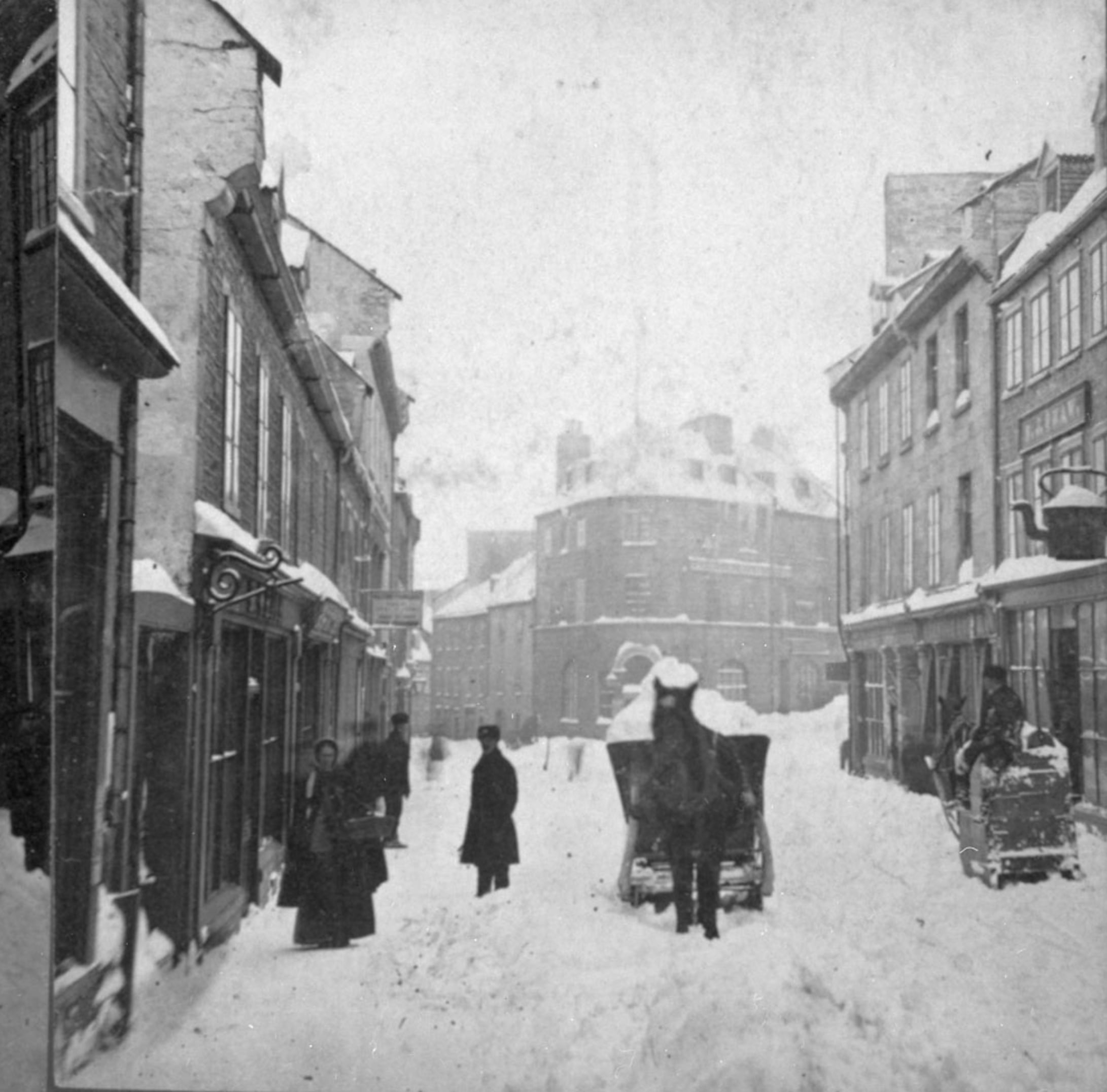 A black-and-white photo of a snowy street lined with old buildings, people in winter clothing walking, and a horse-drawn carriage in the snow. The scene appears to be from the late 19th or early 20th century.