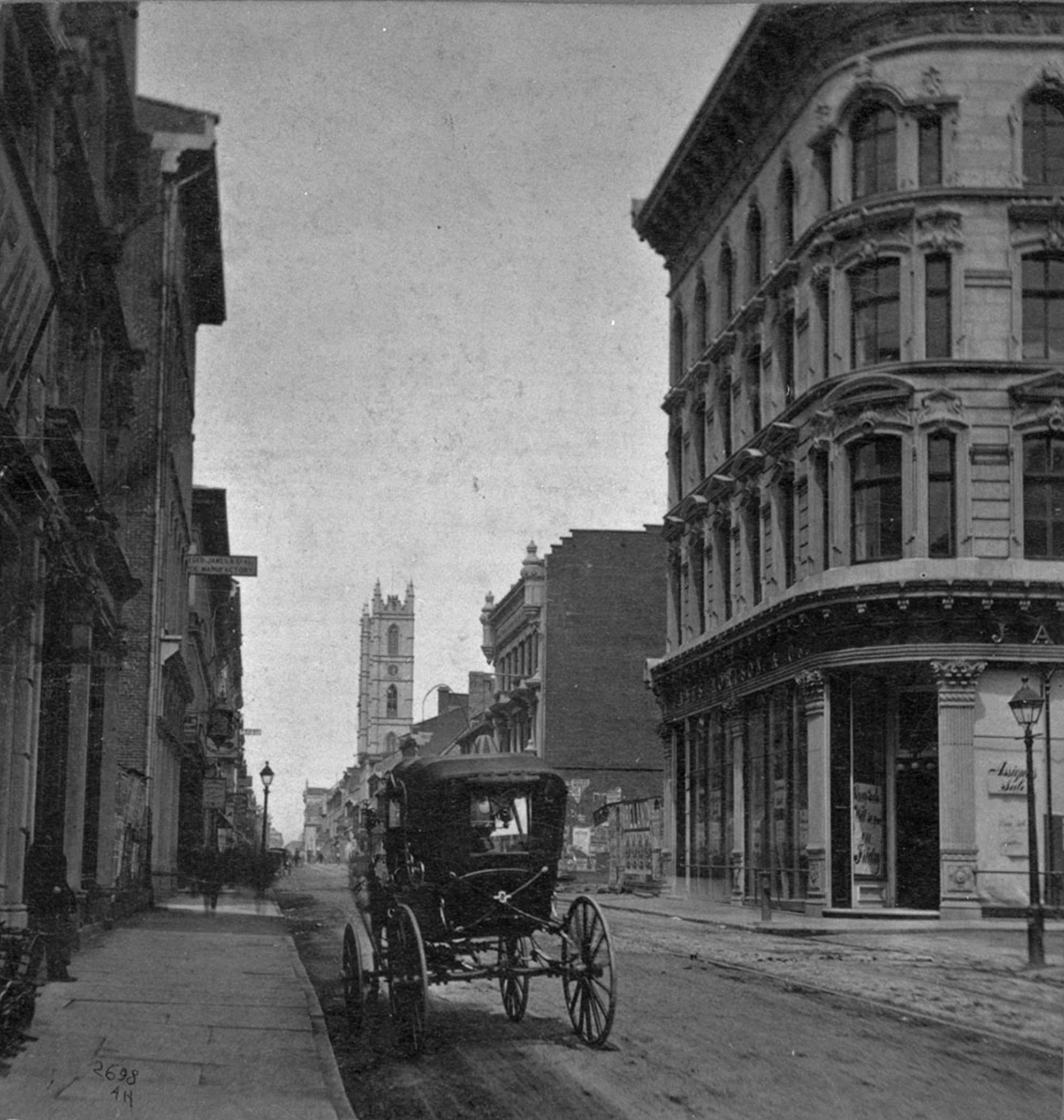 Black and white photo of a horse-drawn carriage on a wide city street lined with tall, ornate buildings; a church with a tall tower is visible in the background.