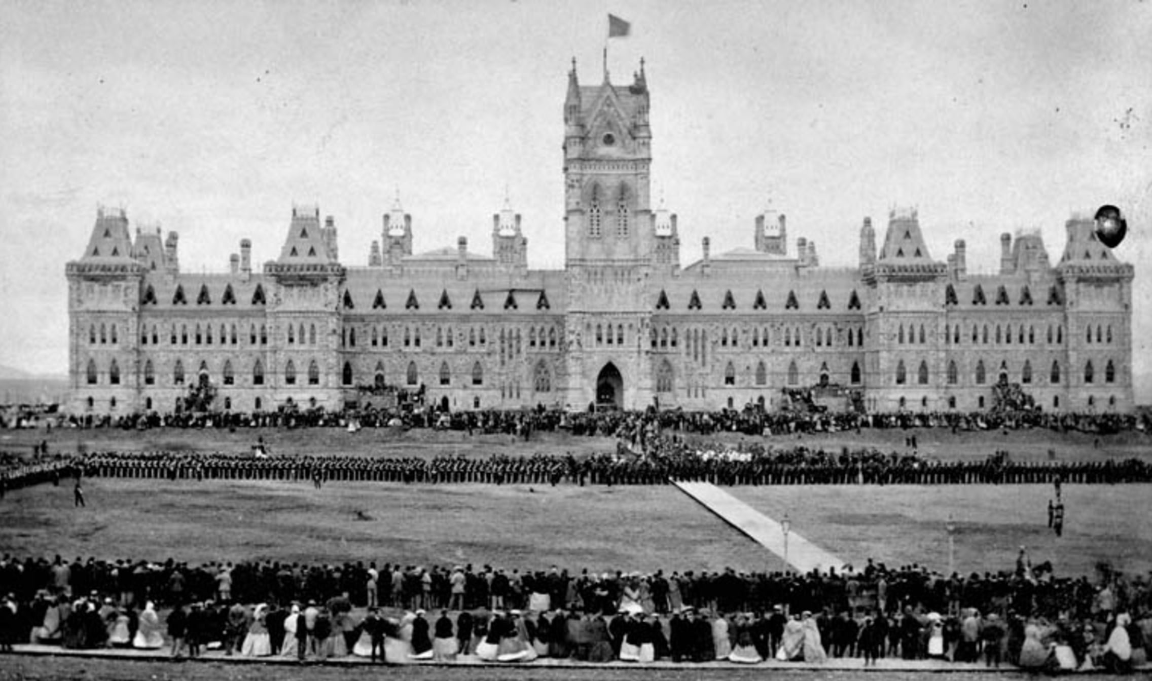 A large crowd gathers in front of a grand, historic Gothic-style building with a central clock tower, wide lawns, and decorative stonework, resembling a government or parliamentary structure.