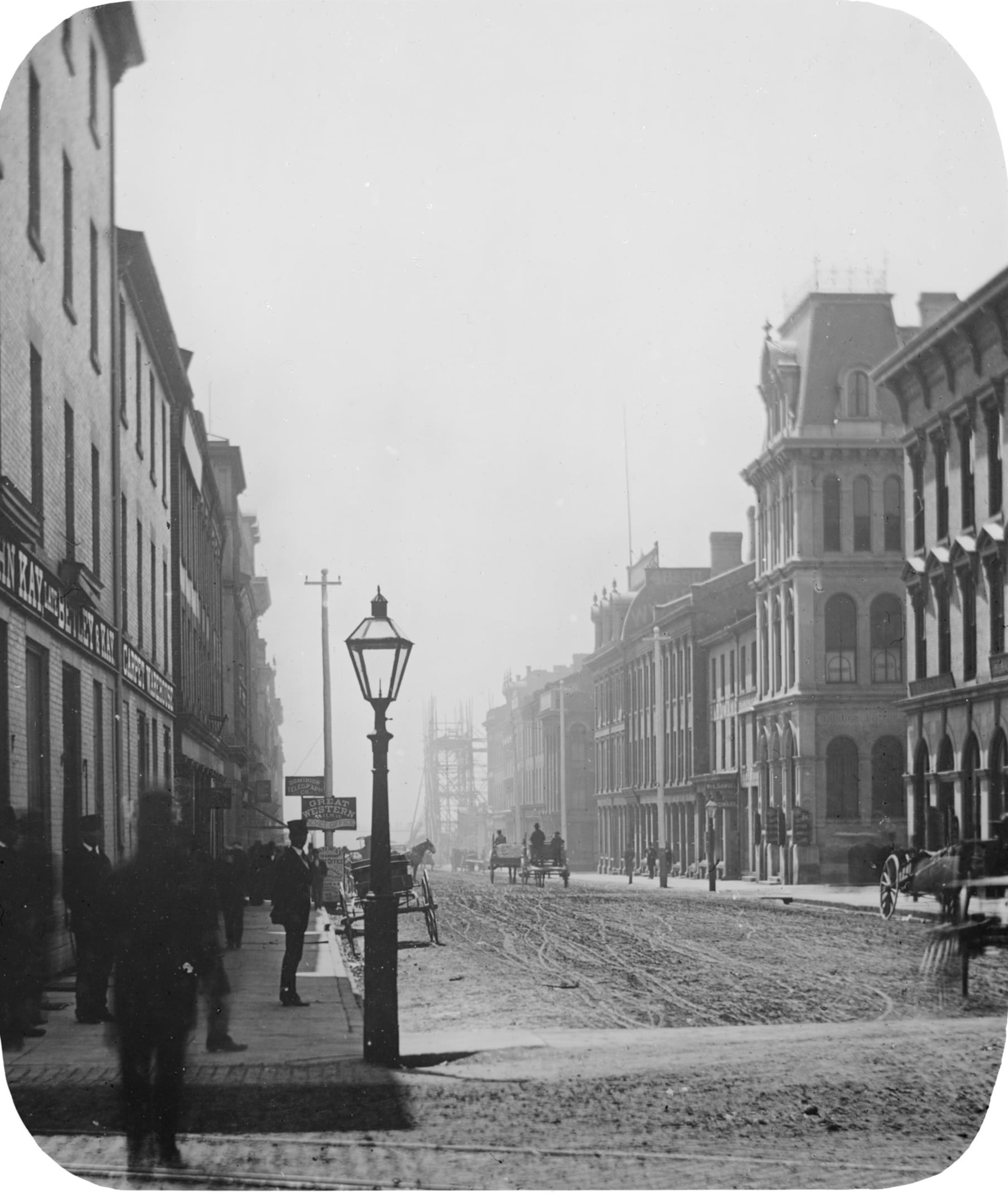 Black and white photo of a city street in the late 1800s, with horse-drawn carriages, people walking on sidewalks, and tall buildings lining both sides. A gas streetlamp stands in the foreground.