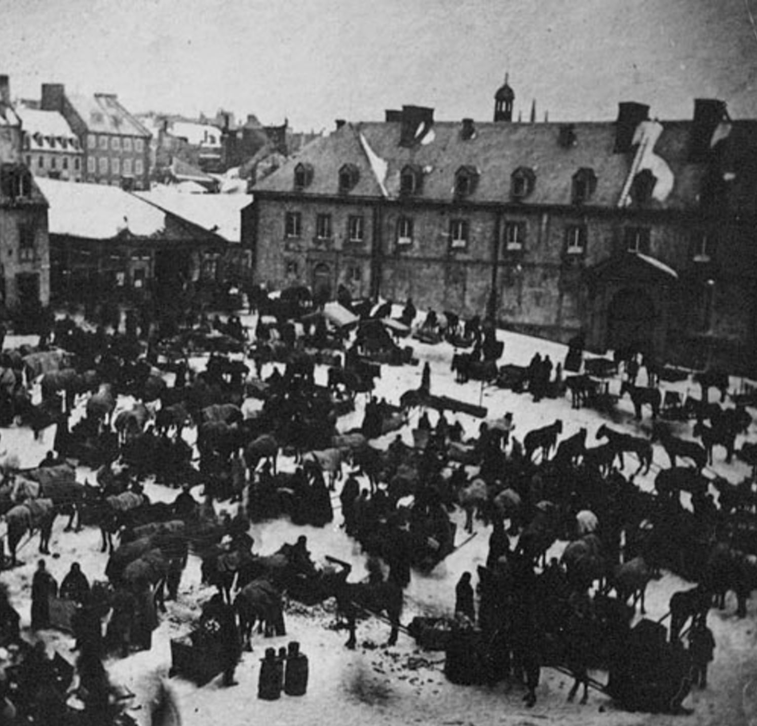 A crowded town square in winter, filled with people and horses on snow-covered ground, surrounded by old stone buildings under a cloudy sky.