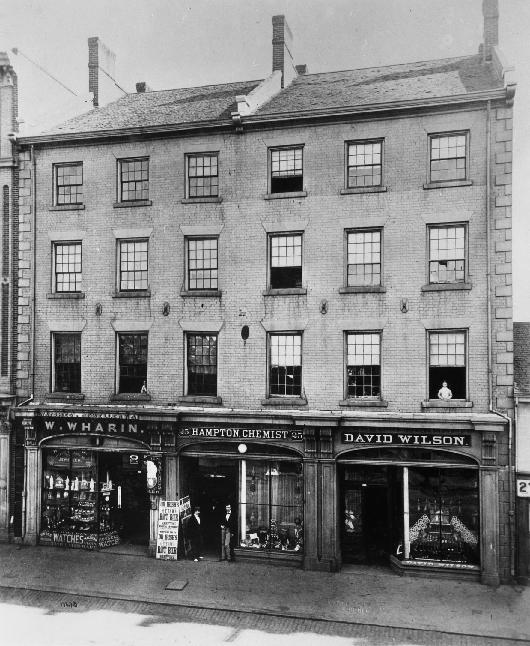 Black-and-white photo of a three-story brick building with storefronts: W. Wharin, Hampton Chemist, and David Wilson. People stand outside the shops, and various goods are displayed in the windows.