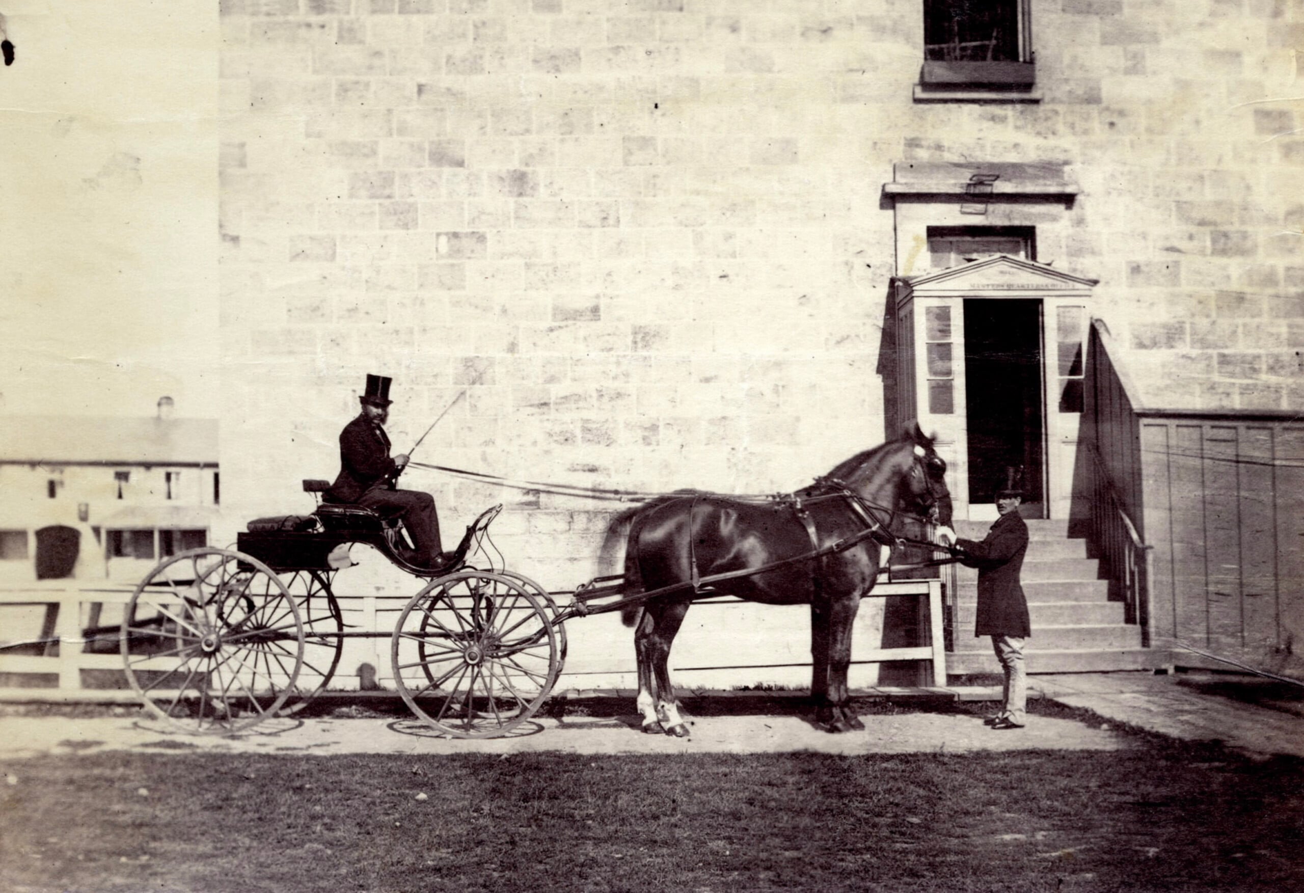 A man in a top hat sits in a horse-drawn carriage while another man stands beside the horse in front of a large stone building with steps leading to a door.