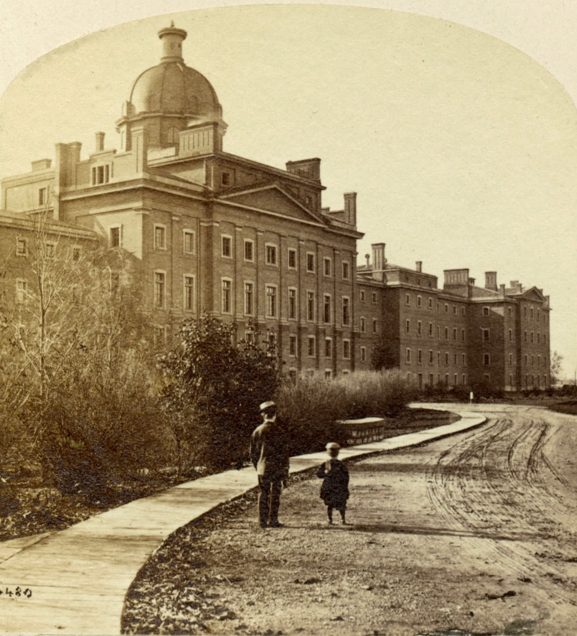 A sepia-toned historical photograph shows an adult and a child standing on a curved walkway in front of a large, multi-story building with a prominent dome and many windows. The area is surrounded by trees and grass.