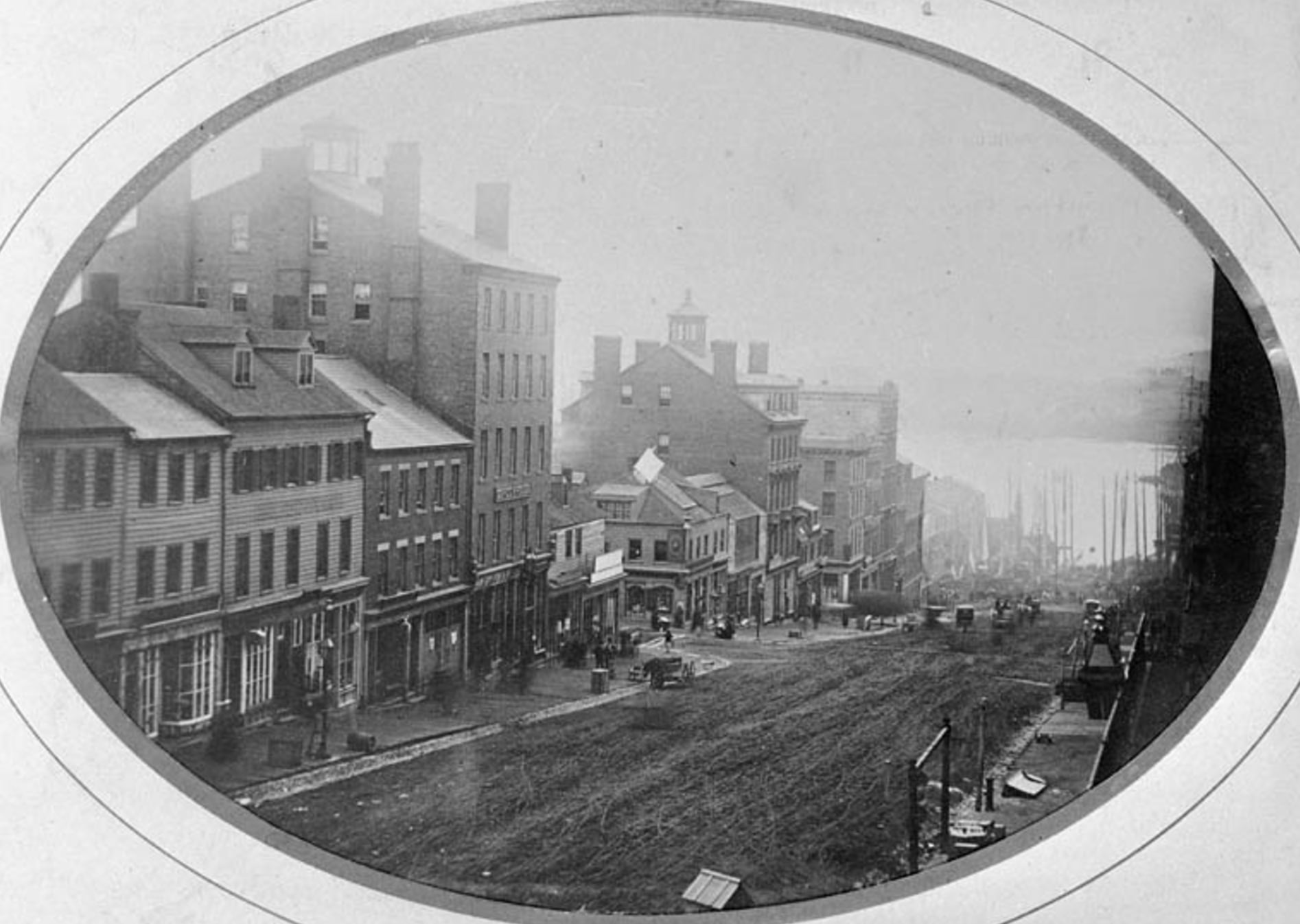 Black and white photo of a 19th-century street with multi-story brick and wooden buildings, people walking, horse-drawn carriages, and a view of a river with docked ships in the background.