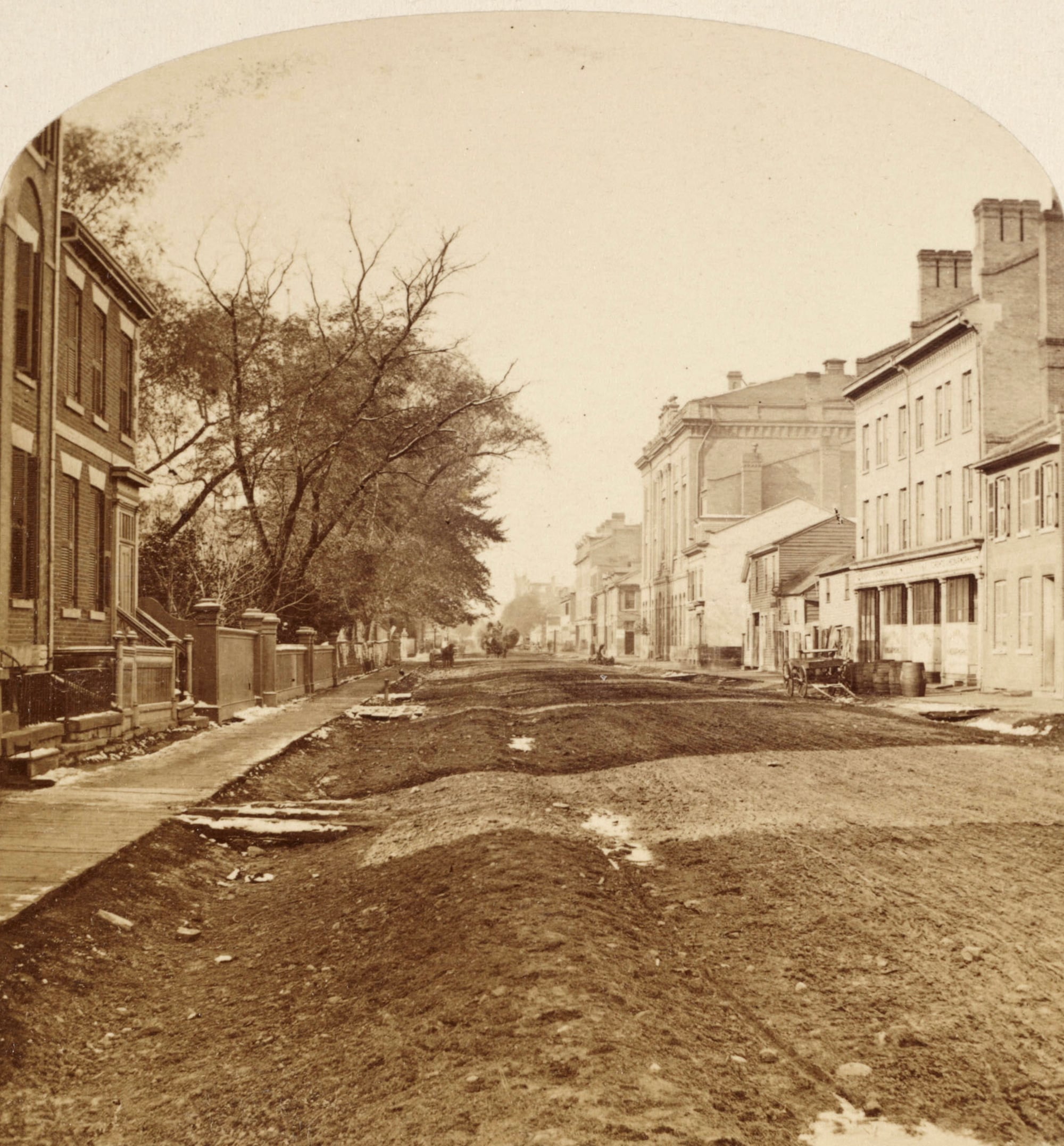 Sepia photo of a 19th-century city street with dirt roads under construction, lined with trees and brick buildings, including houses and larger structures, with debris scattered along the road.