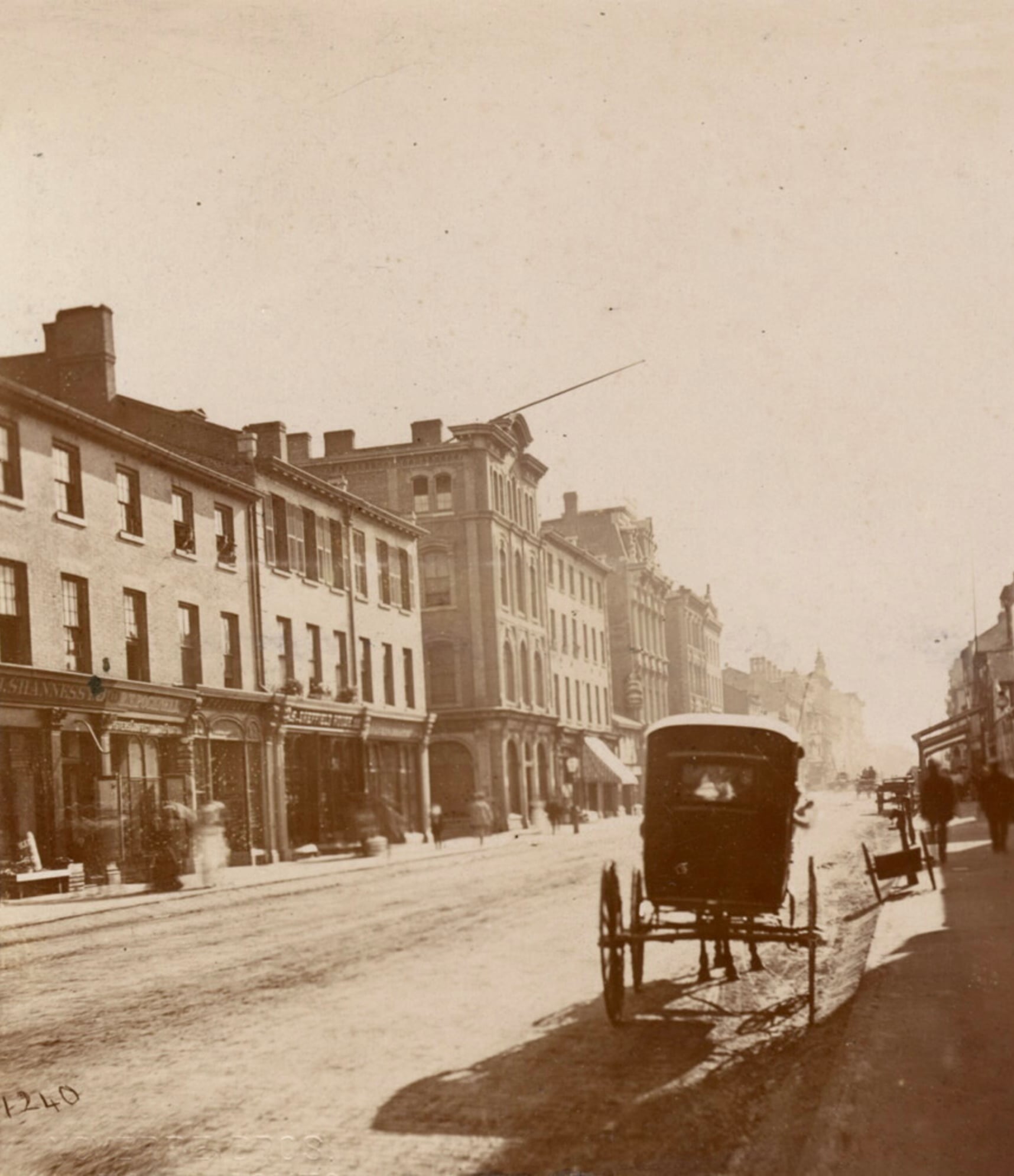 Sepia photo of a city street lined with brick buildings, shops, and businesses. A horse-drawn carriage stands by the sidewalk, while people walk along the street. The scene suggests a late 19th or early 20th century setting.