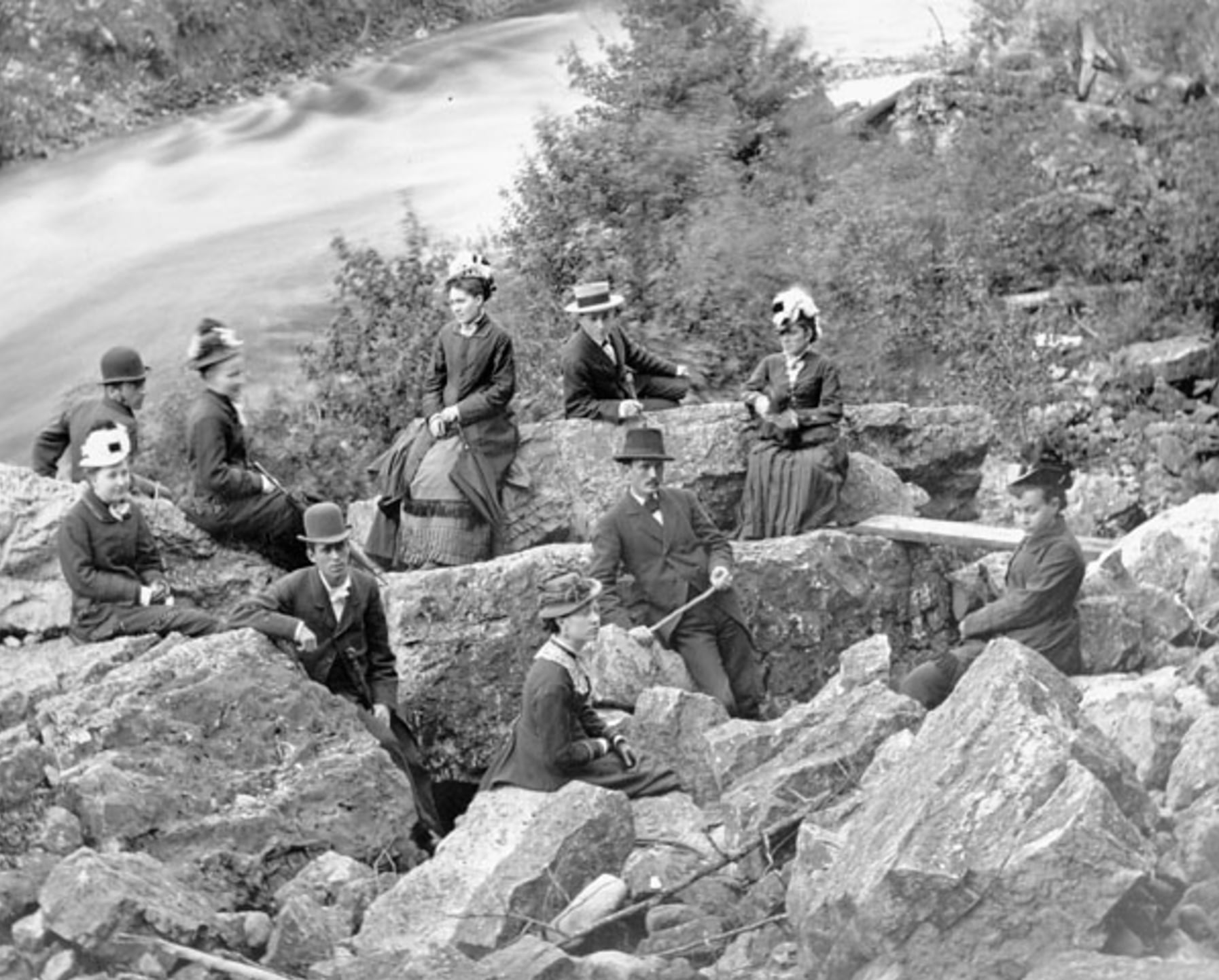 A group of men and women in 19th-century clothing sit and pose on large rocks beside a river, surrounded by trees and vegetation.