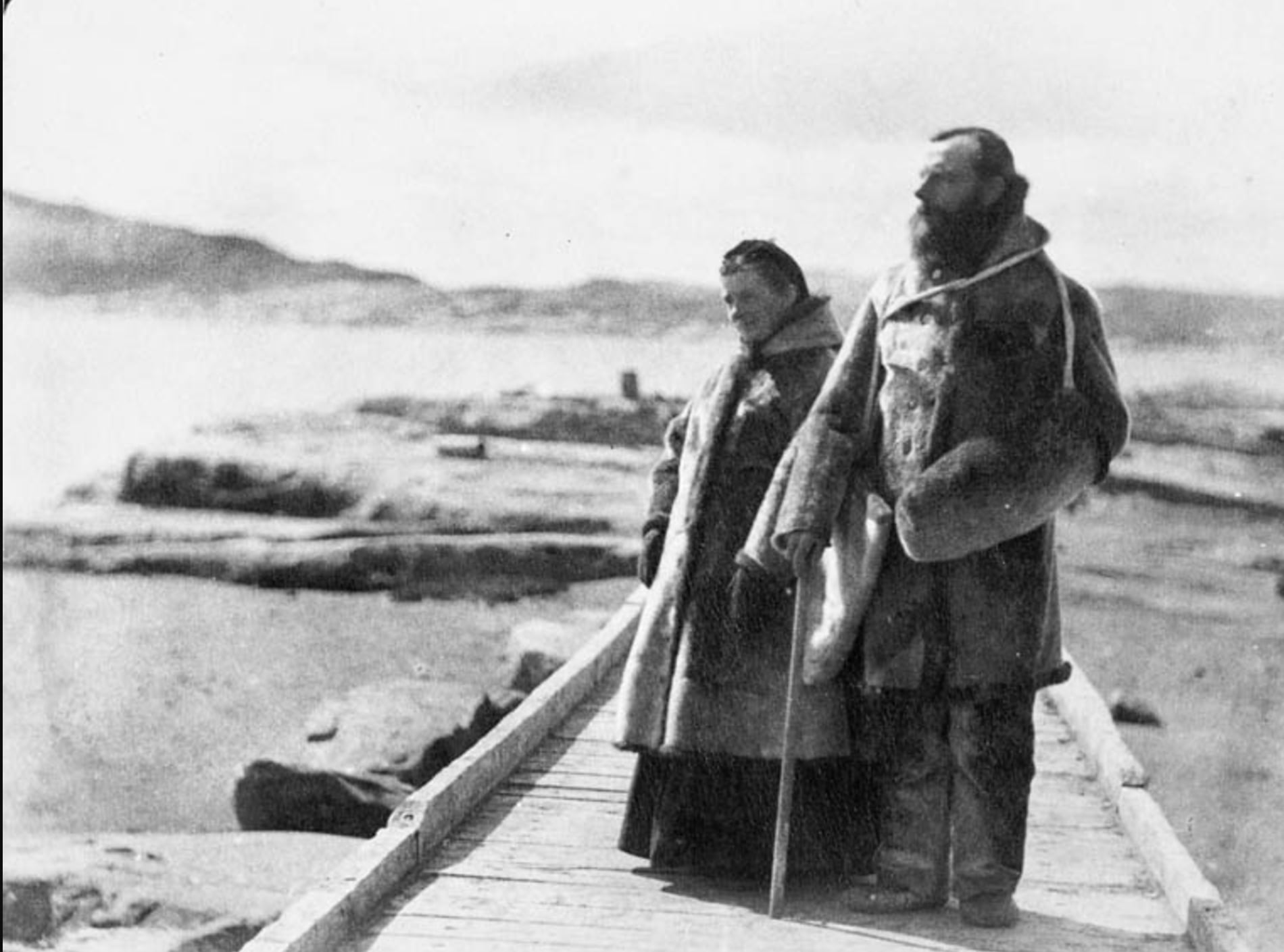 A man and a woman in heavy fur clothing stand on a wooden walkway by a rocky, icy shoreline, with mountains and water in the background. The man holds a staff and both face toward the distant landscape.