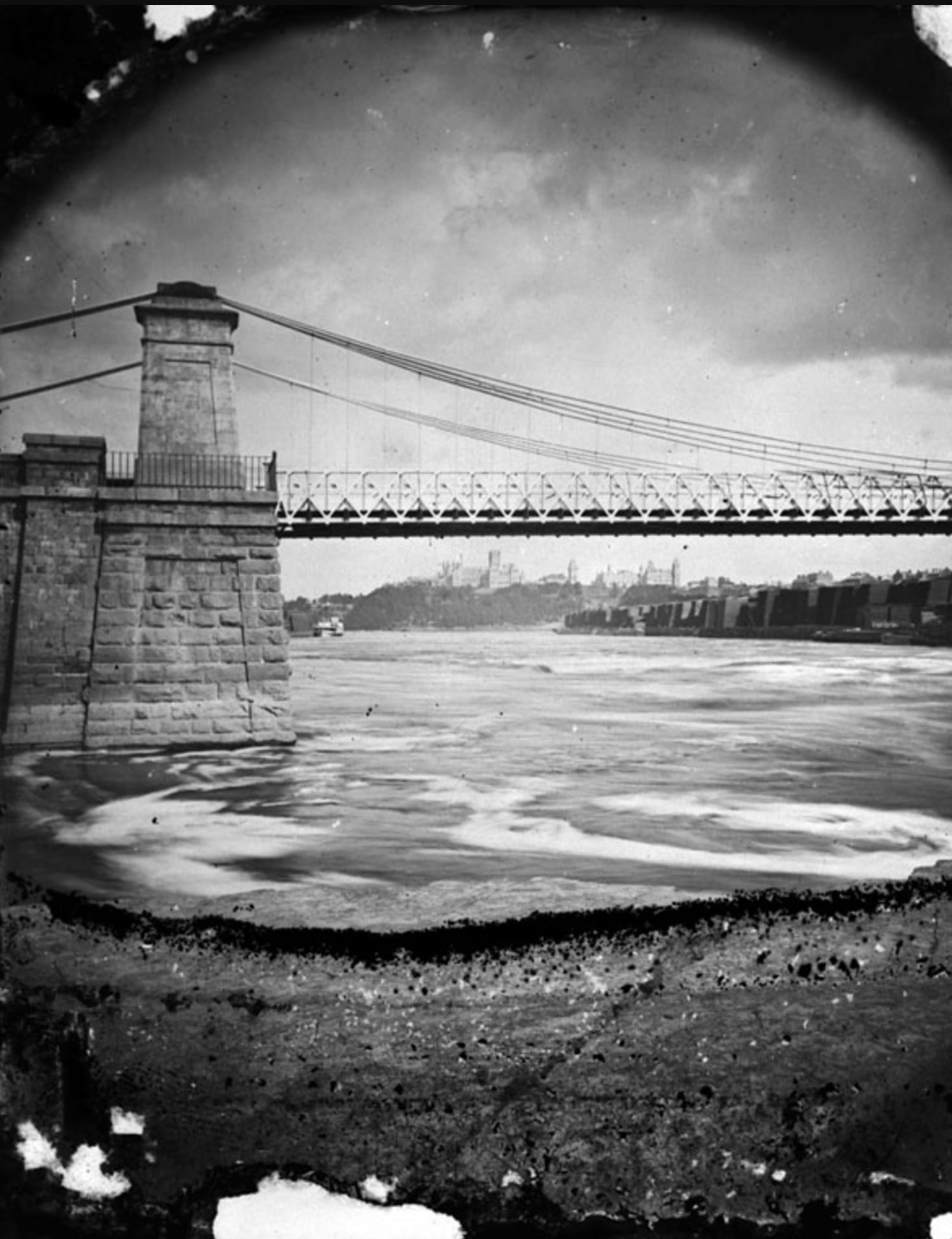 A black-and-white photo shows a suspension bridge crossing a wide river, with stone supports and churning water below. Buildings and trees are visible in the distance under a cloudy sky. The photo edges appear weathered or damaged.