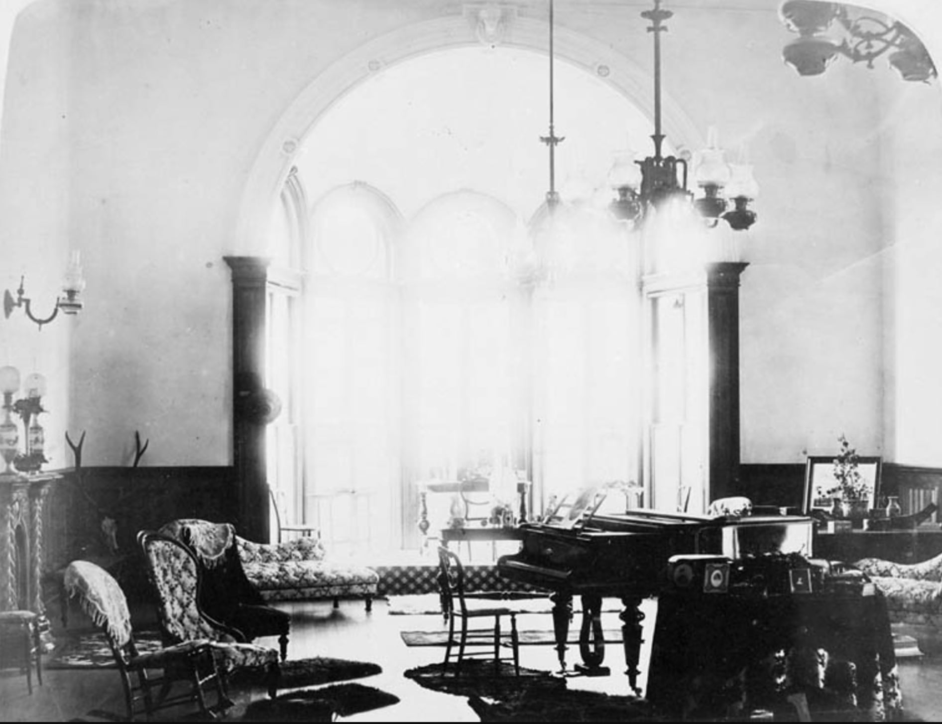 Black and white photo of an elegant Victorian-era parlor with arched windows, a grand piano, ornate chairs, sofas, a chandelier, and various decorative furnishings. Sunlight streams through the large windows.