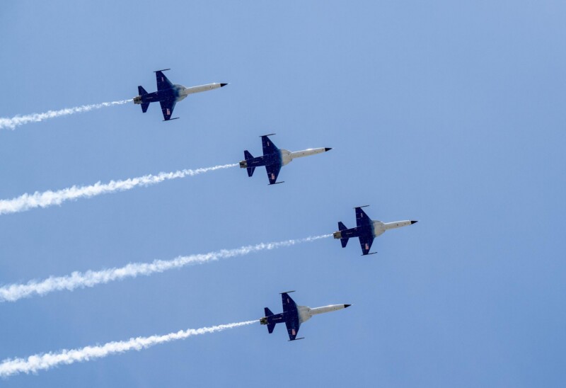 Four fighter jets fly in a tight formation against a clear blue sky, leaving white vapor trails behind them.