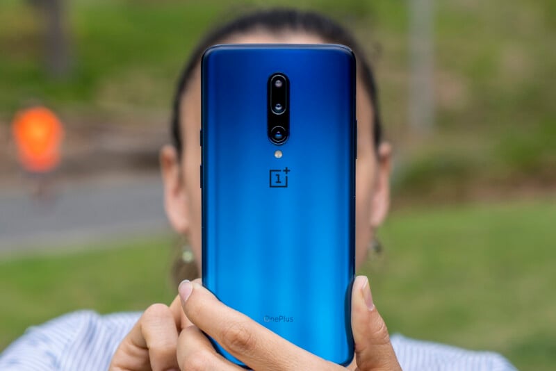 A woman holds a smartphone toward the camera. The phone has a blue gradient design. 