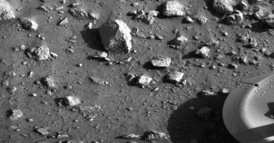 A rocky, dusty surface with scattered stones of various sizes. Part of a circular, metallic object appears in the lower right corner. The scene looks barren and textured.