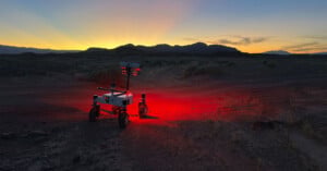 A small rover with lights glowing red underneath stands on rugged terrain at dusk, with mountains silhouetted against a colorful sunset sky in the background.