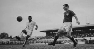 A black-and-white photo shows two soccer players on a field, one kicking the ball mid-air as the other approaches. Spectators fill the stands in the background.