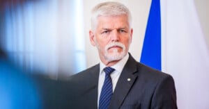 A man with white hair and a beard wearing a gray suit, white shirt, and blue striped tie stands in front of a blue and white flag, looking serious.