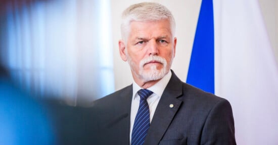 A man with white hair and a beard wearing a gray suit, white shirt, and blue striped tie stands in front of a blue and white flag, looking serious.